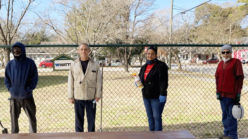 Two men and two women standing in front of a black fence with a red picnic table in front of them. 
