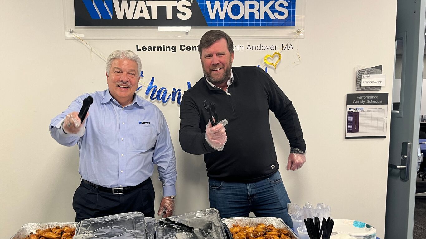 Two men standing in front of a table of food with utensils ready to serve food for Watts Employee Appreciation Day.