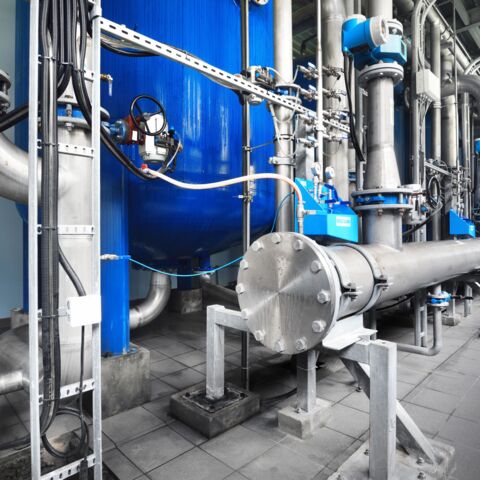 Data center water treatment system featuring large stainless-steel pipes, valves, instrumentation, and a blue pressure vessel. Automated control devices and sensors are mounted throughout the piping network, with the system installed on a clean tiled floor inside a data center